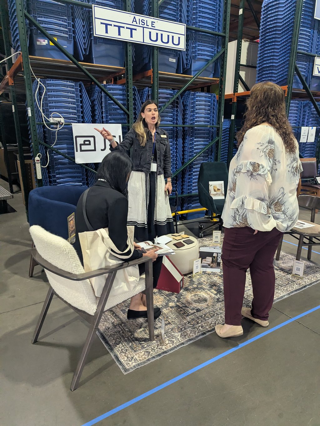 Three attendees talking around a small furniture display in an aisle of blue bin racking