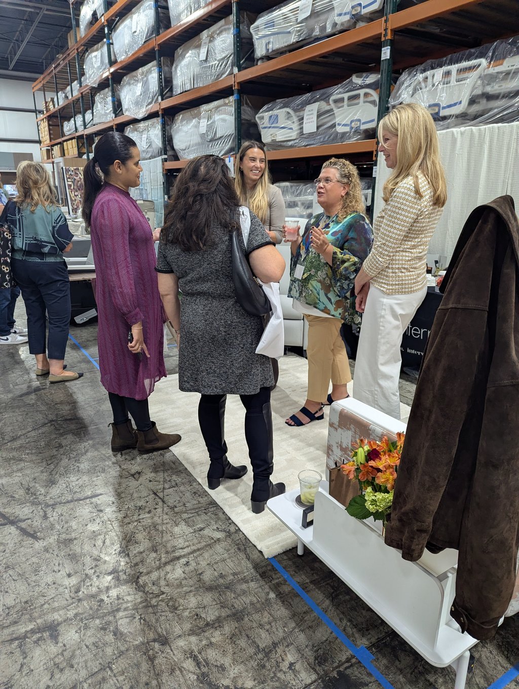 A small group of attendees chatting with drinks in front of racks of wrapped mattresses and bed frames