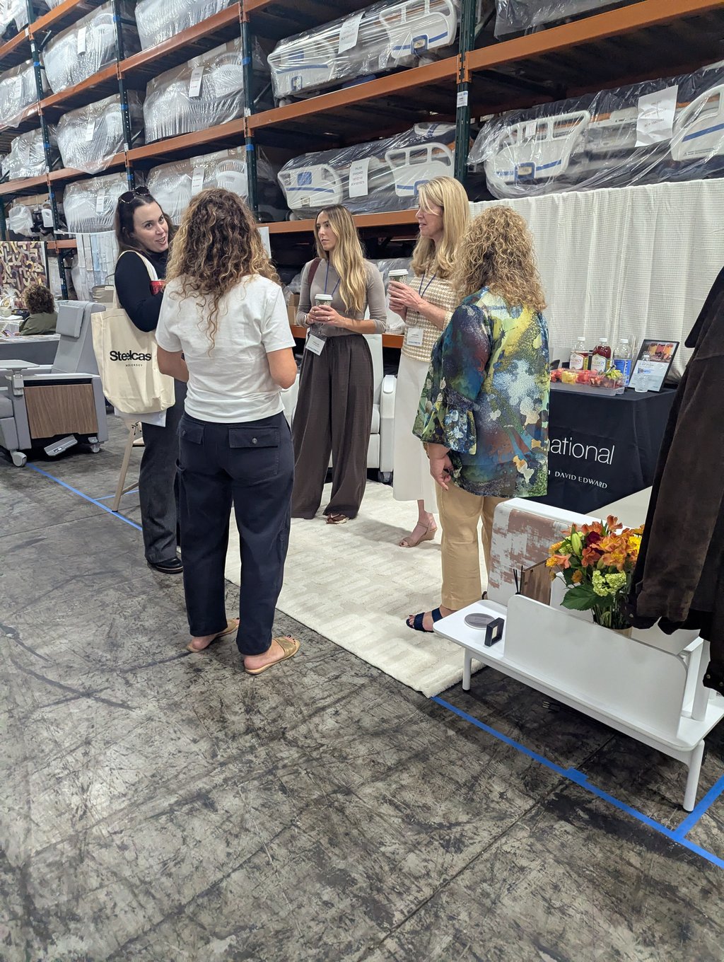 Five attendees with coffee cups talking in a showroom aisle in front of wrapped beds and mattresses
