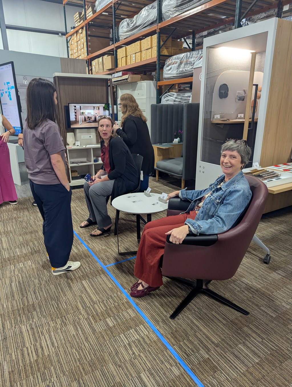 Four attendees talking among seating displays and a sample table beside a white room panel