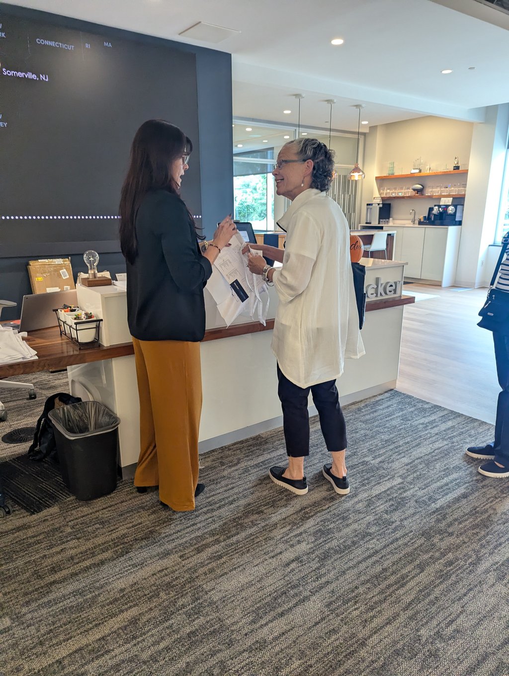 Two attendees talking at a reception counter in a bright office beside a wall screen listing locations