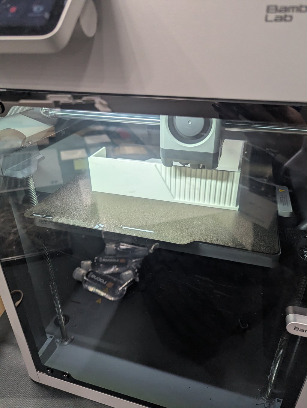 A white ribbed part being printed on the build plate inside a Bambu Lab 3D printer