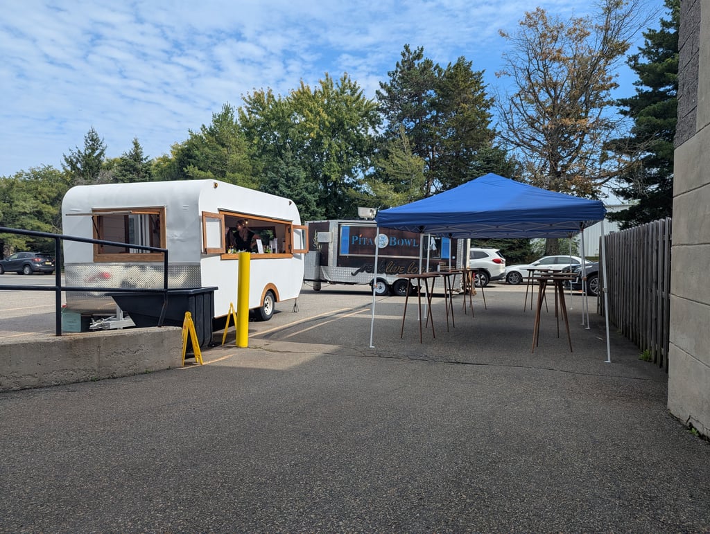 Two food trailers and a blue canopy with picnic tables under trees in the car park
