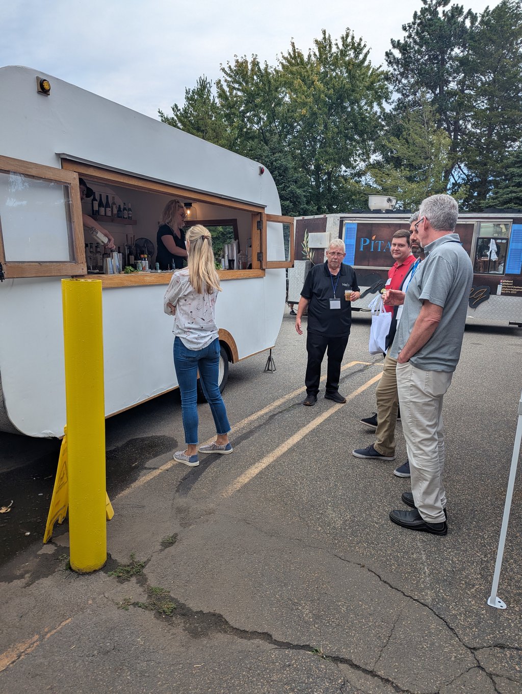 Attendees gathered at the window of a white mobile bar trailer with a food truck behind