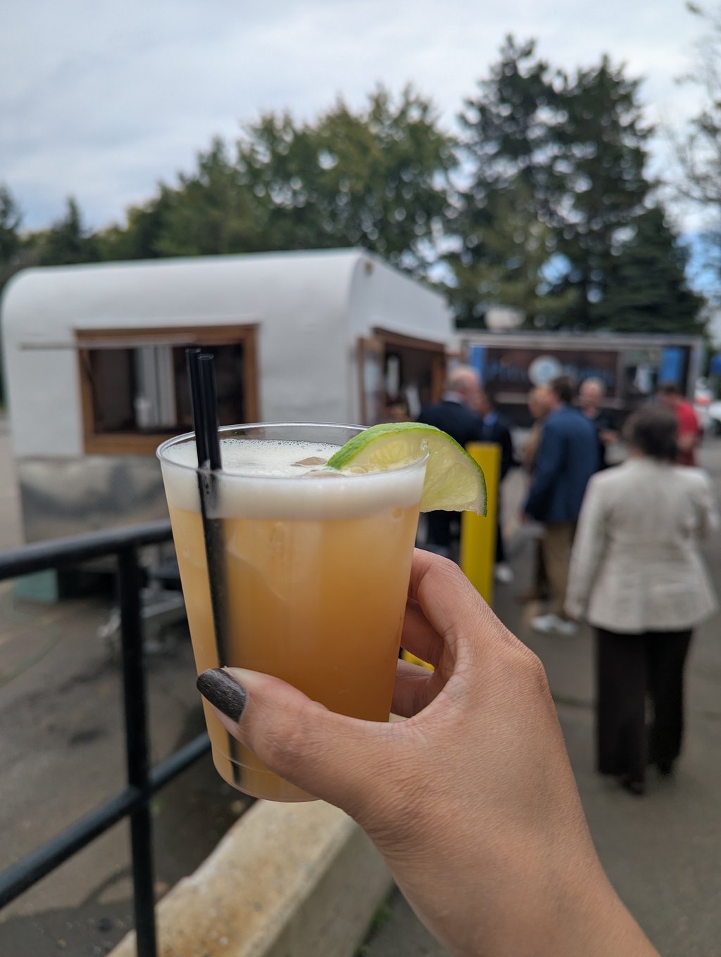 A hand holding a cup of frothy pale drink with two straws and a lime wedge, food trucks blurred behind