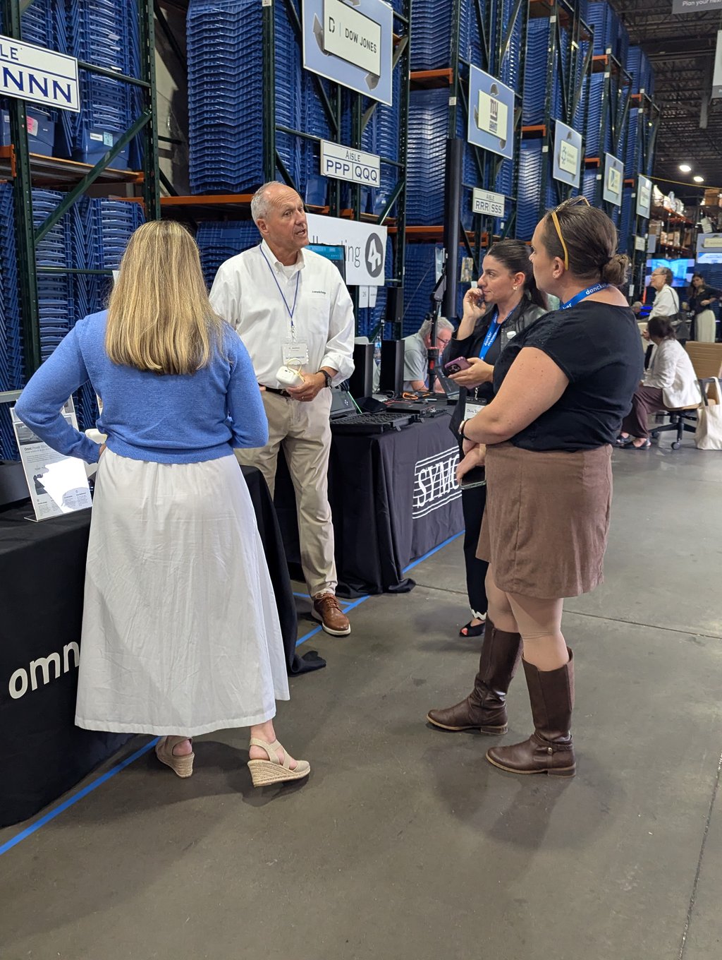 Four attendees talking at a display table in an aisle of blue bin racking by the Dow Jones sign