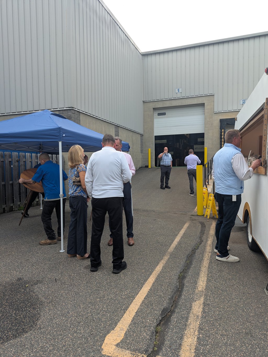 Attendees standing outside the grey industrial building by an open loading door, canopy and food truck nearby