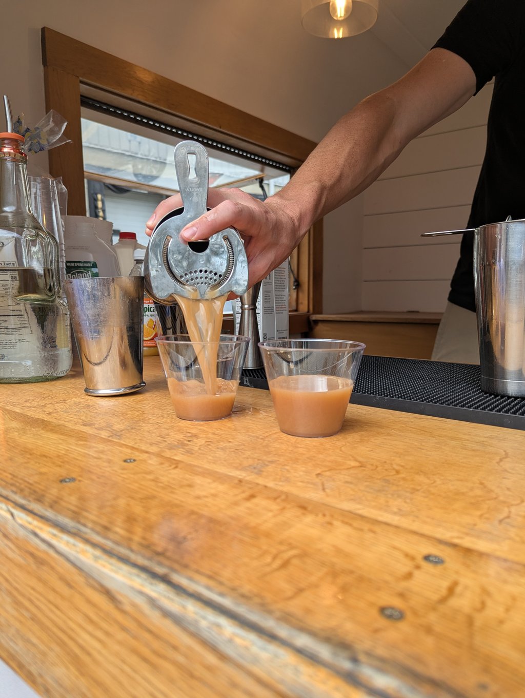 A bartender straining a tan cocktail from a shaker into two cups on a wooden counter