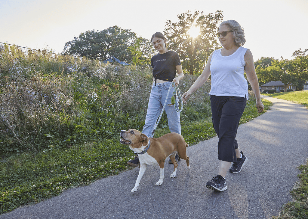 Two women walking a dog on a paved park path beside wildflowers at sunset