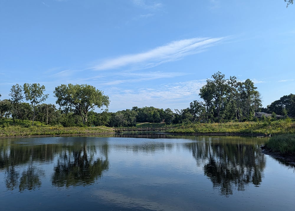 Calm pond reflecting trees and blue sky with a wooden footbridge in the distance