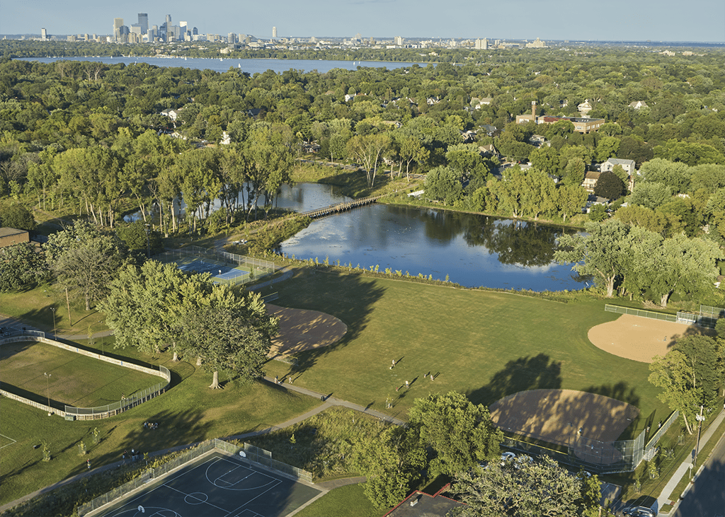 Aerial view of Morningside park with baseball diamonds, tennis courts, a pond with footbridge, and city skyline in the distance