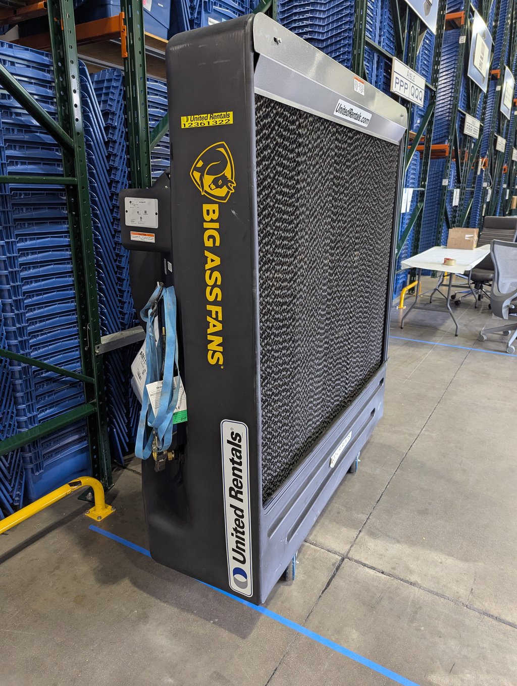 A large industrial floor fan standing beside racked storage bins during setup