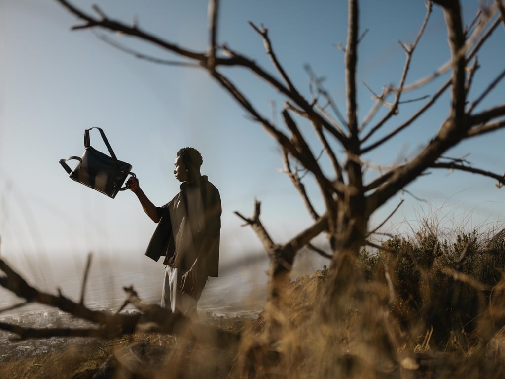 Silhouette of a person holding a backpack aloft on a windswept hillside against a pale blue sky