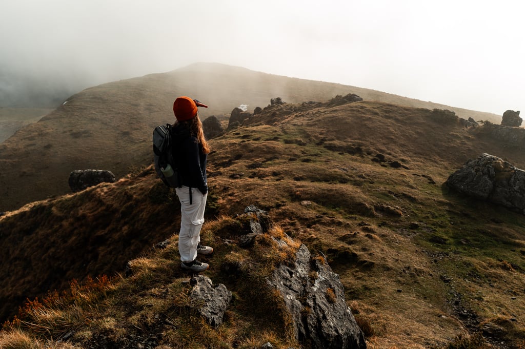 Hiker in an orange beanie standing on a rocky outcrop overlooking fog-shrouded hills