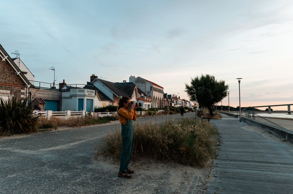 Person in green overalls photographing on a coastal promenade with dunes and a bridge in the distance
