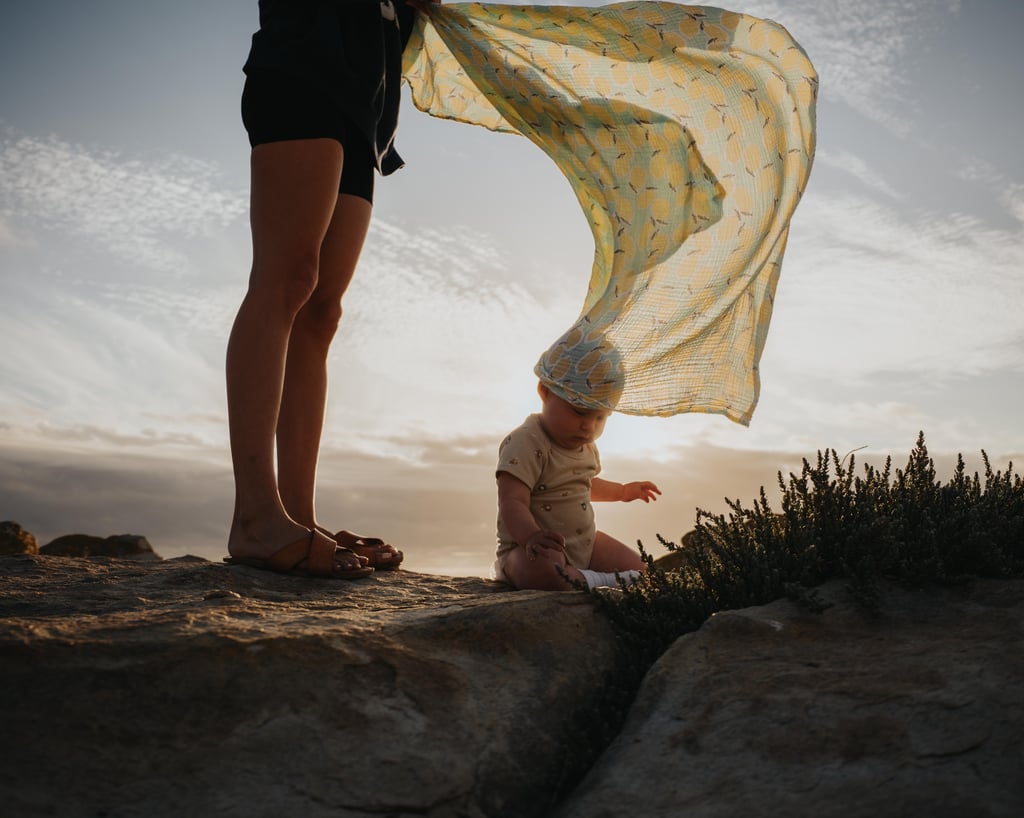 Sunset scene on a rocky ledge with an adult standing beside a seated baby, cloth billowing overhead