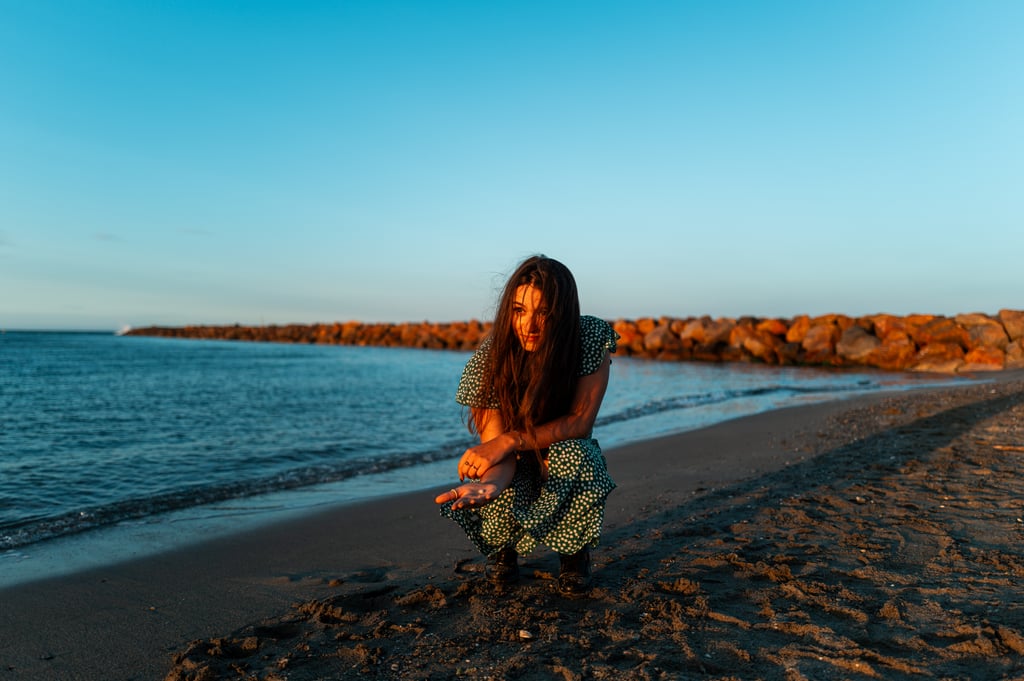 Person crouched on a sandy beach near the water at sunset wearing a green patterned dress