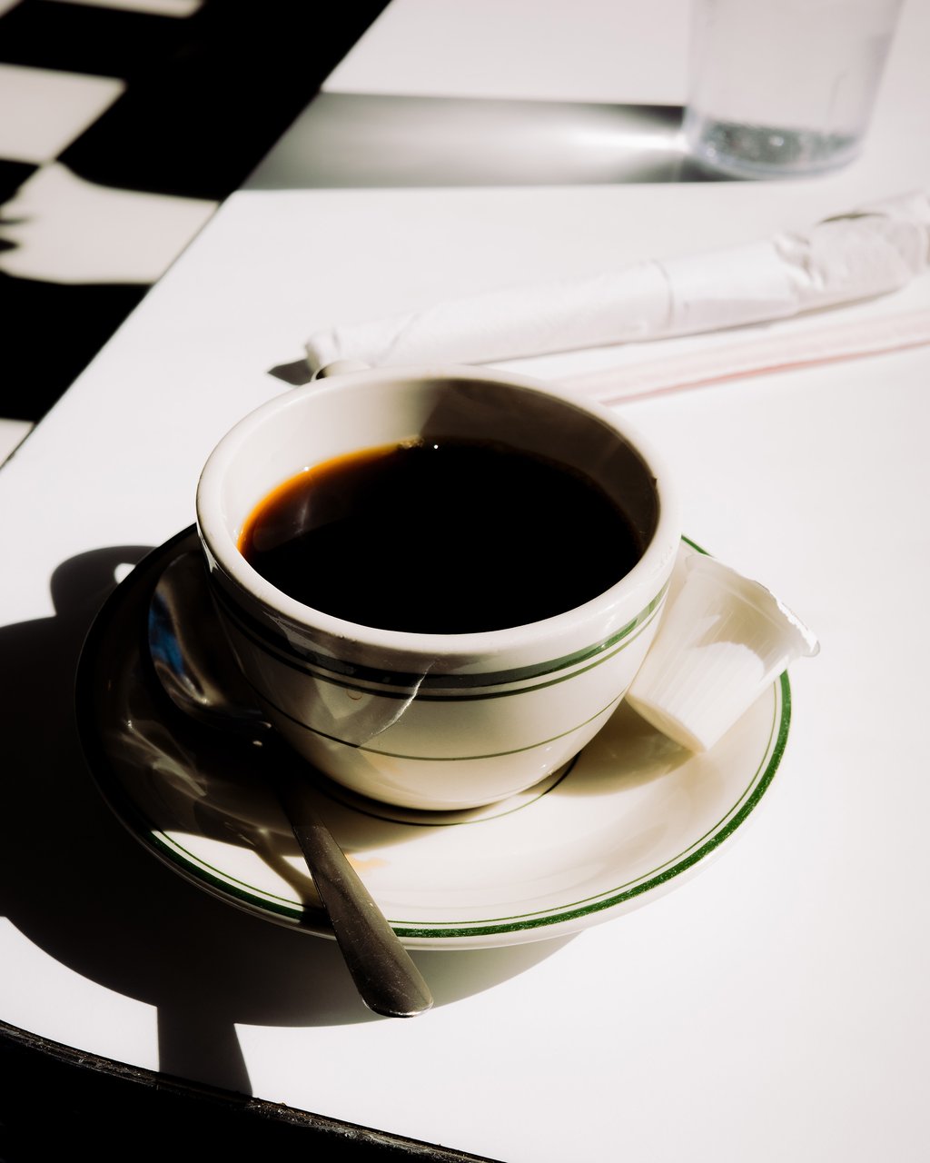 White ceramic cup of black coffee on a saucer with bright sunlight casting strong shadows on a white tabletop
