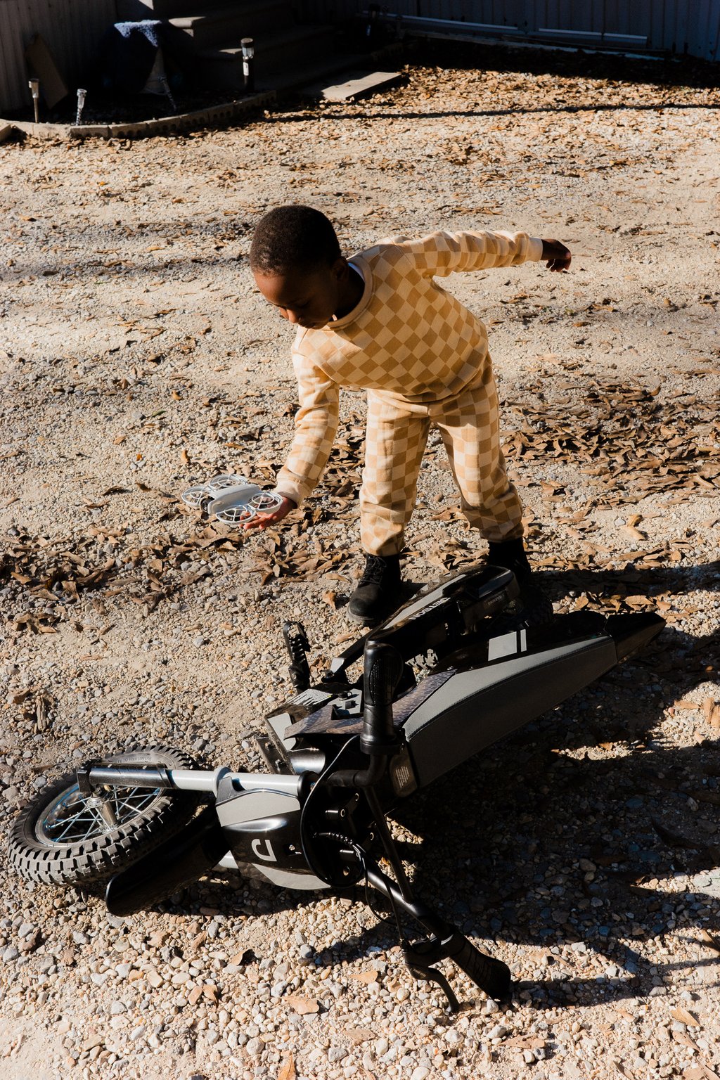 Child in a beige checkered outfit bending over on a gravel driveway to pick up a small white drone