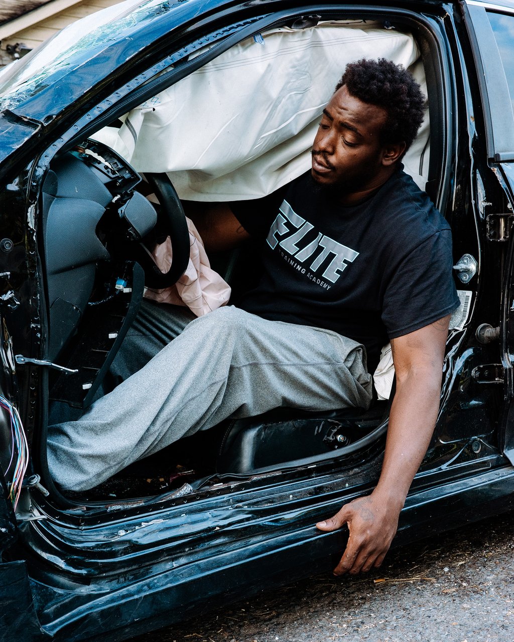 Man sitting inside a heavily damaged car with deployed airbags, leaning out of the driver's side door