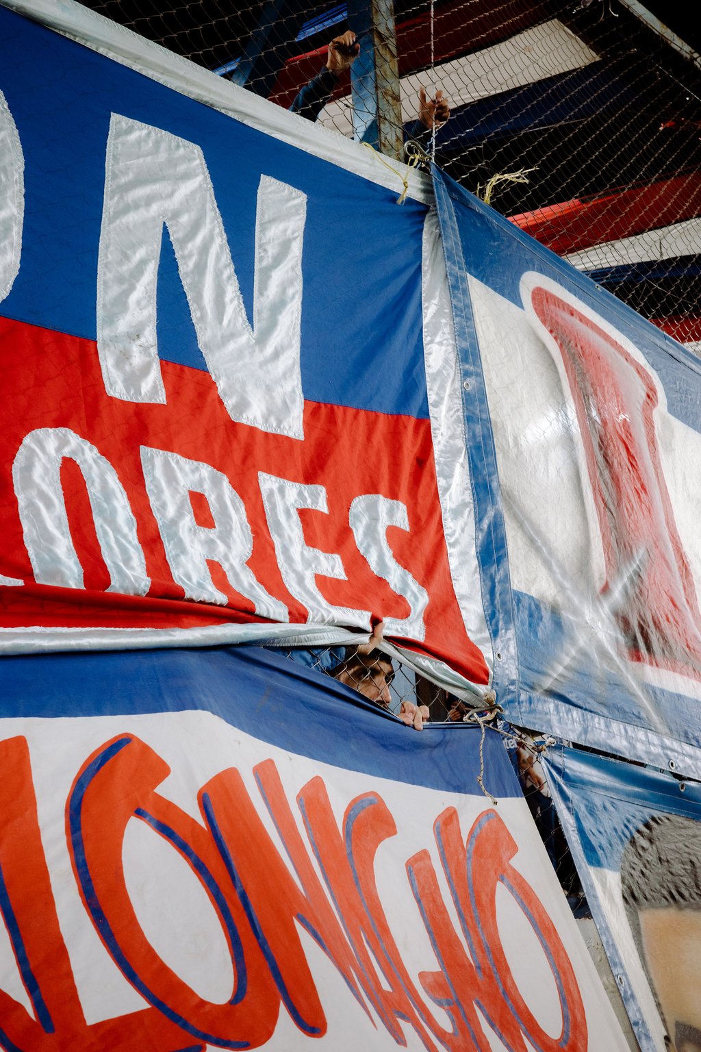 Close-up of layered red, white, and blue banners with a large white letter N and hands gripping chain-link fencing behind