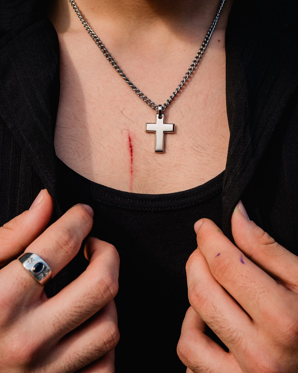 Close-up of a person's chest wearing a silver cross pendant on a chain with hands pulling open a black shirt