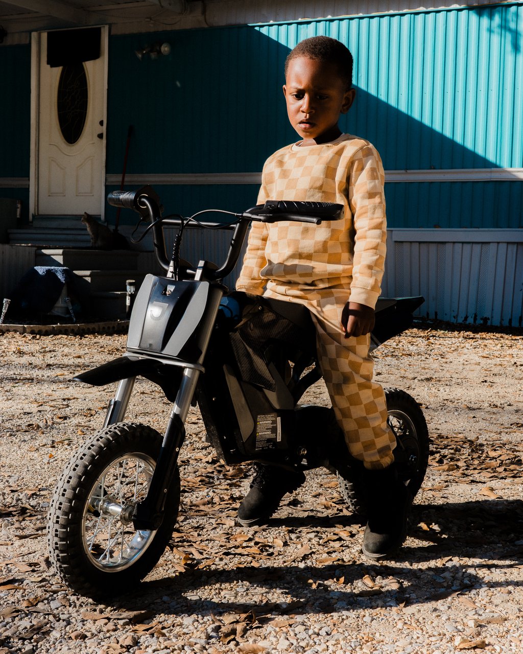 Young boy in a beige checkered outfit sitting on a small black dirt bike in front of a teal mobile home