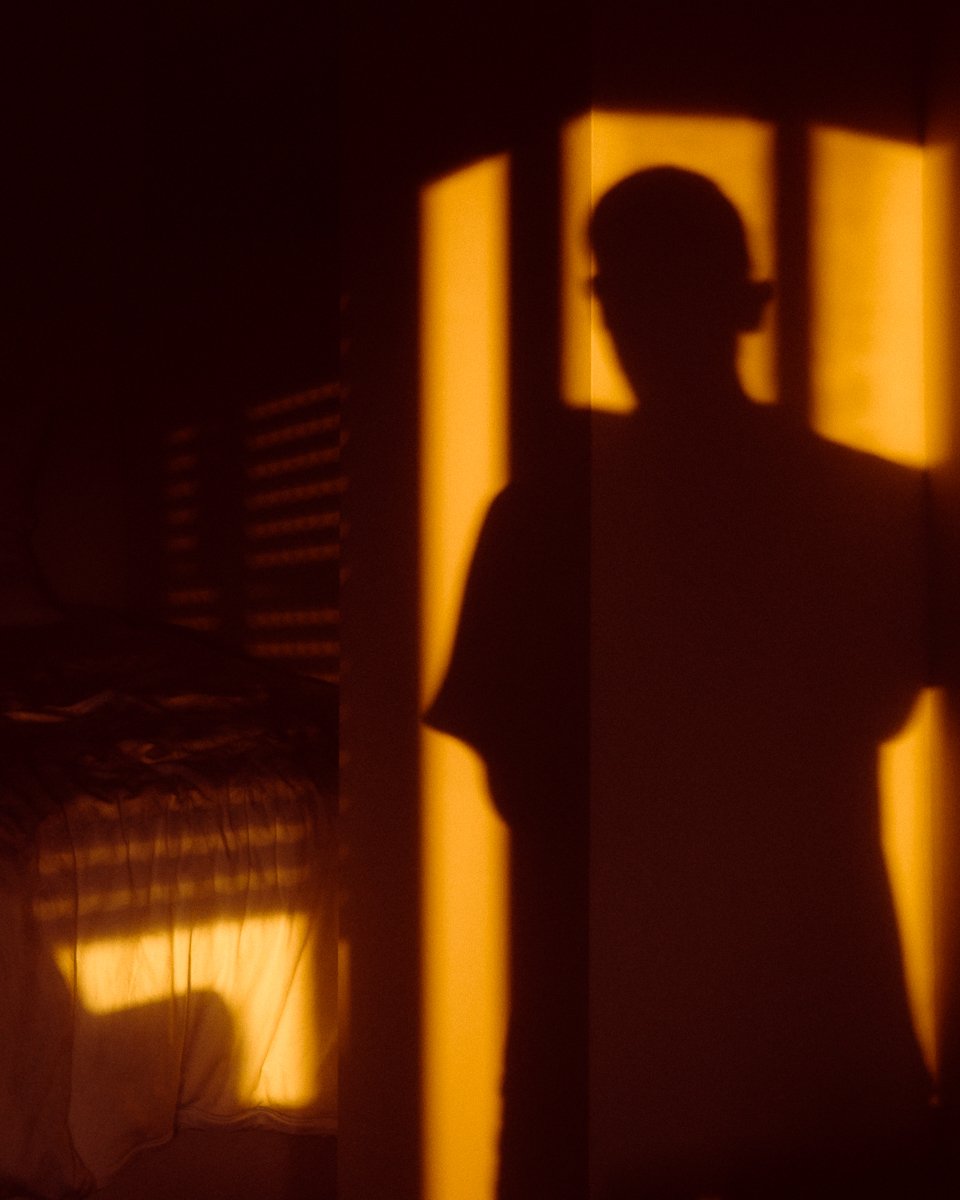 Warm orange sunlight projecting window-pane shadows and a person's silhouette onto an interior wall with rumpled bed sheets