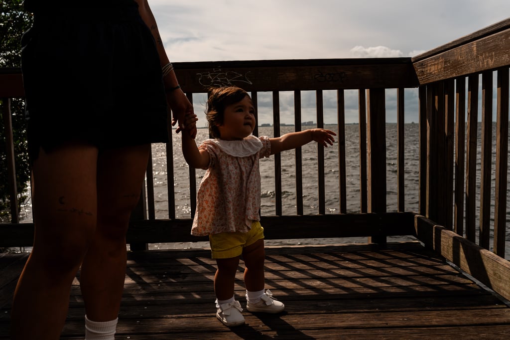 Toddler in a floral top standing on a wooden pier reaching outward while holding an adult's hand