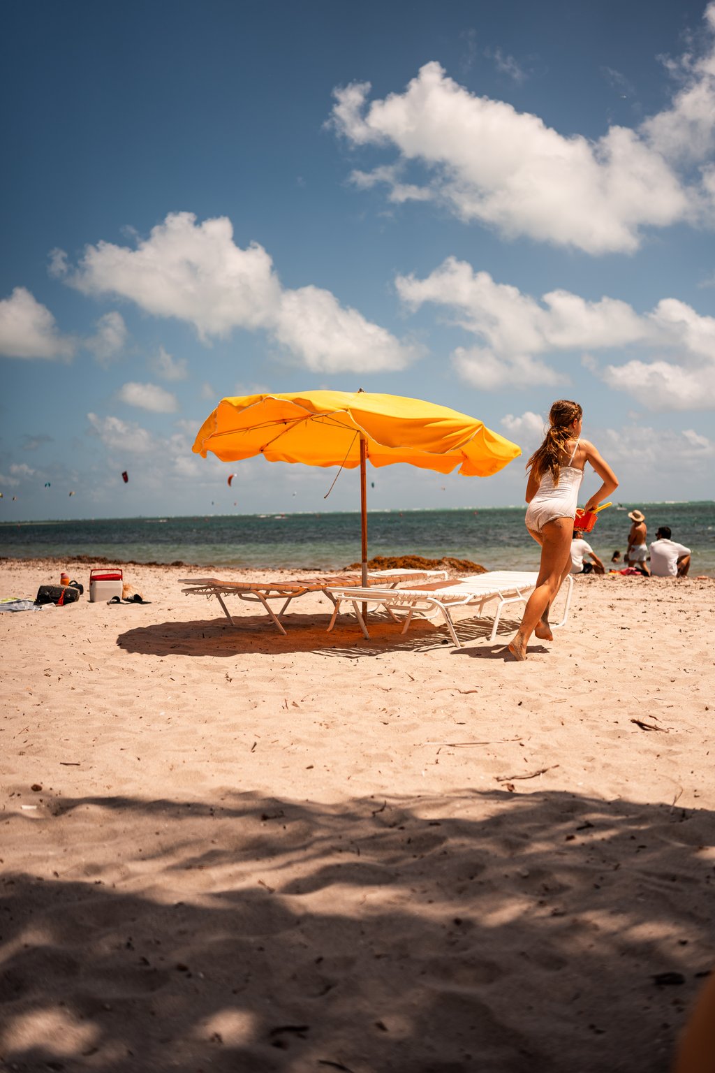 Sunny beach scene with a yellow umbrella over lounge chairs and a woman walking toward calm water