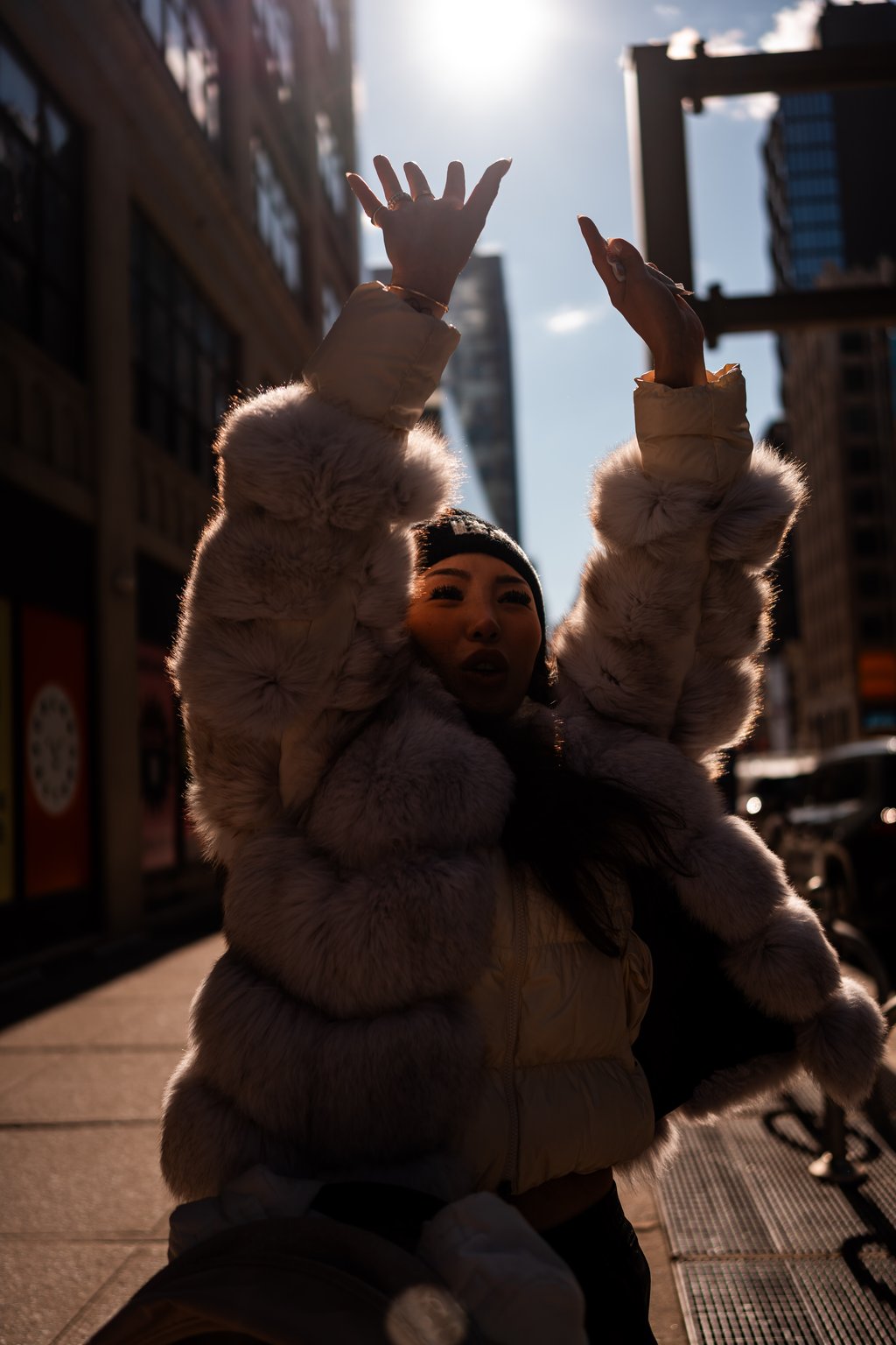 Person in a fur-lined puffer coat and knit hat raising both hands on a sunlit city street