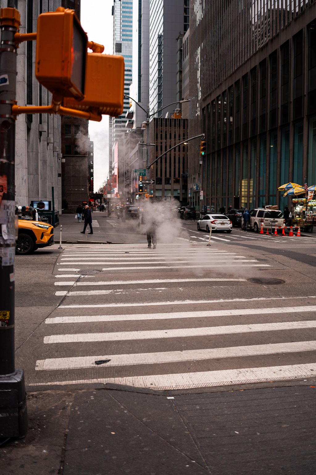 City street with a wide zebra crosswalk and steam rising from a manhole among tall office buildings