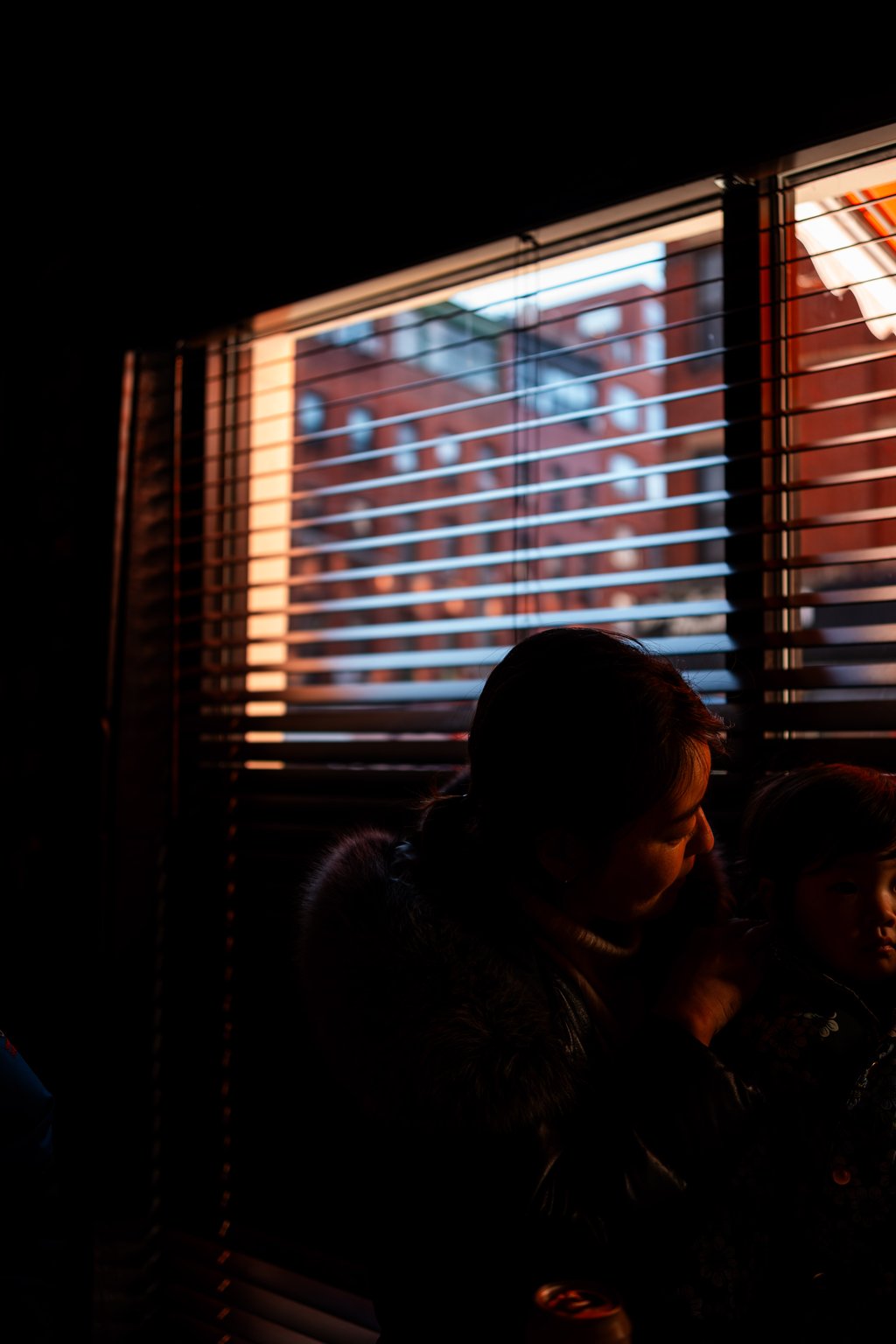 Dim interior with horizontal blinds lit by warm evening light and a silhouetted adult and child