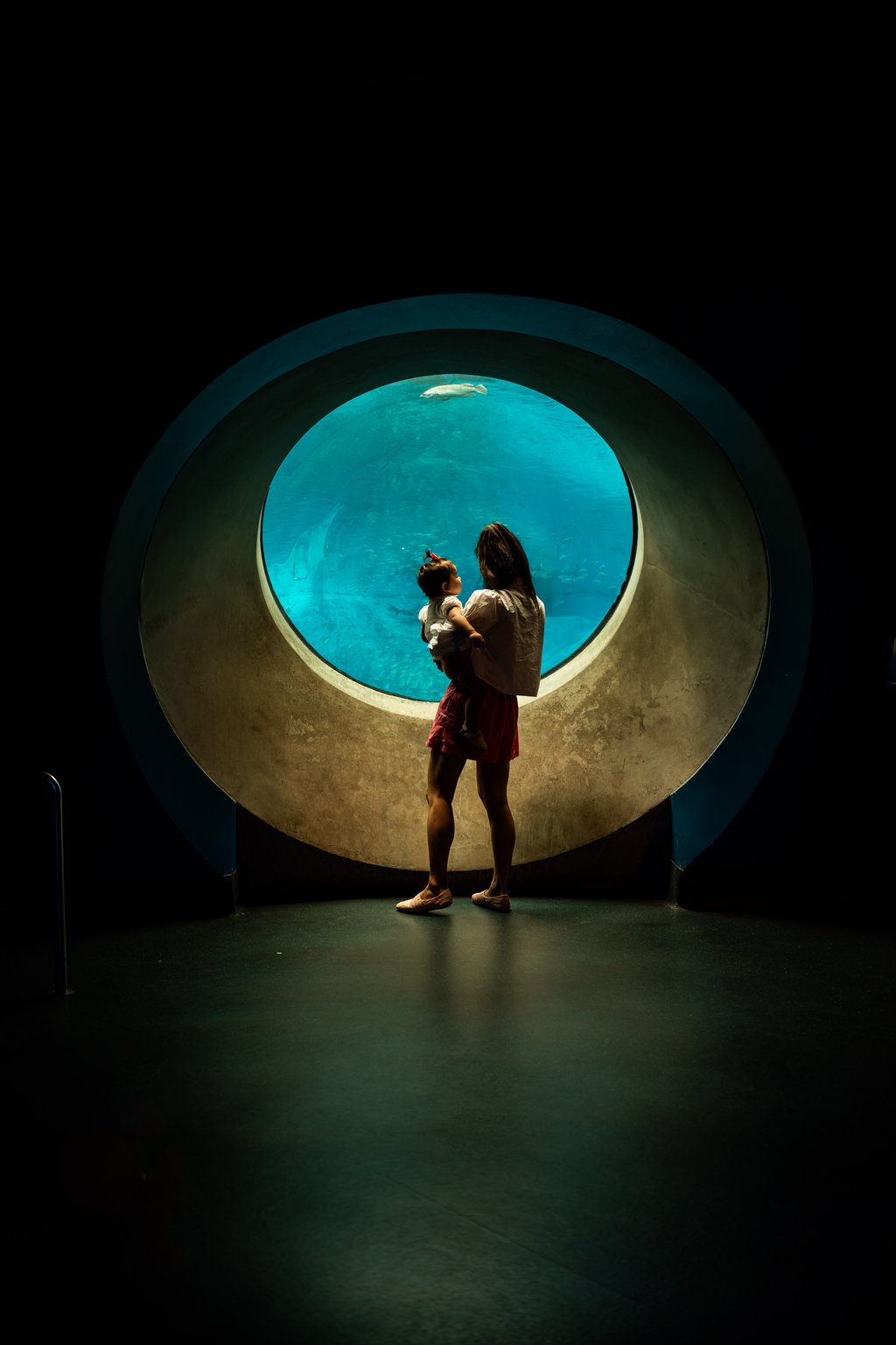 Silhouetted woman holding a toddler before a large circular aquarium porthole with turquoise water