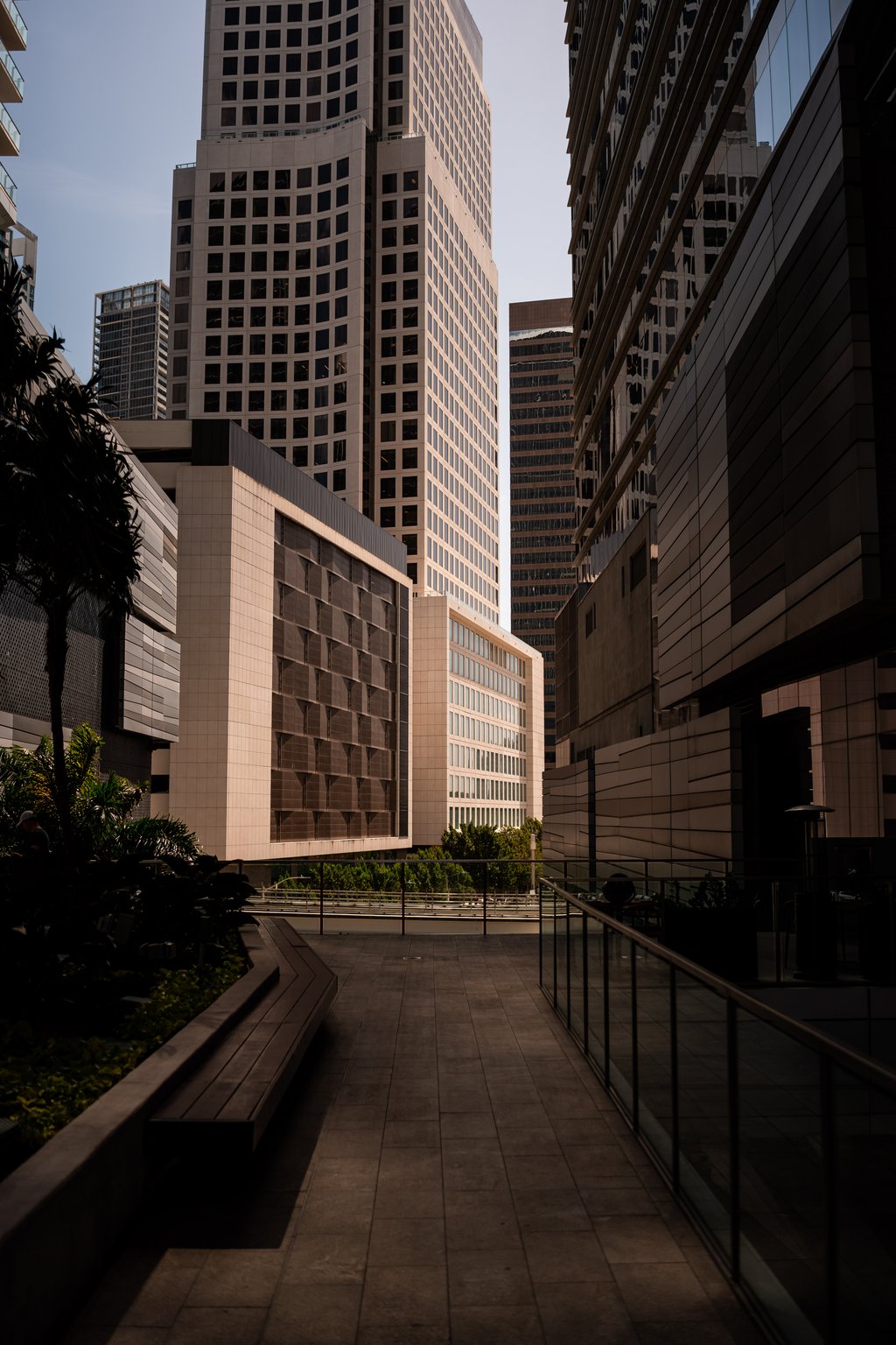 Sunlit urban canyon of tall modern office towers with a shadowed pedestrian walkway and benches