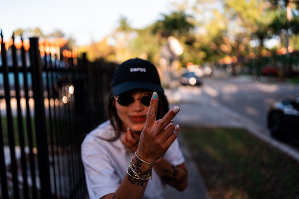 Person in a black cap and sunglasses holding up two fingers toward the camera with bracelets and a wrist tattoo