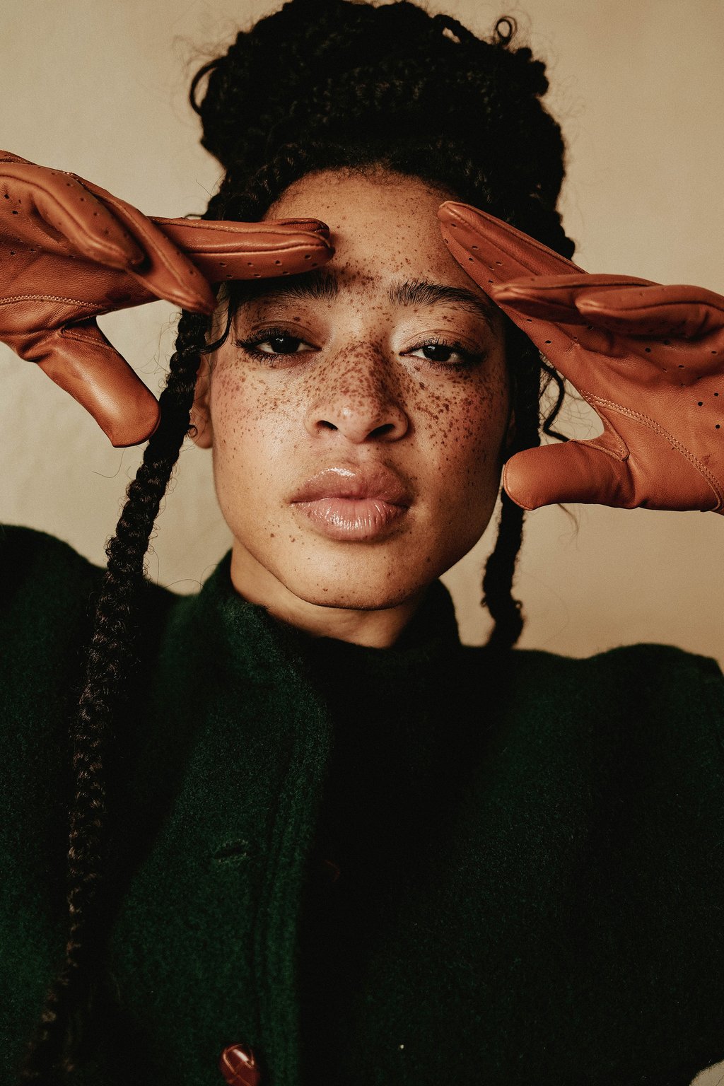 Freckled person with braided hair and glossy lips, brown leather gloves framing their face, wearing a dark green coat