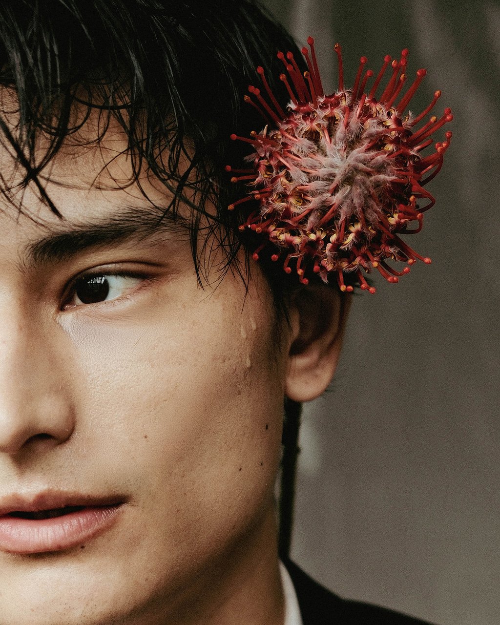 Close-up of a young person's face with dark wet hair and a large round red spiky flower tucked behind the ear