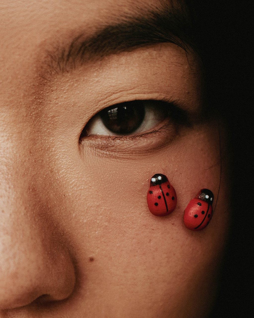 Extreme close-up of a person's eye with visible skin texture and two small red ladybug decorations on the cheek