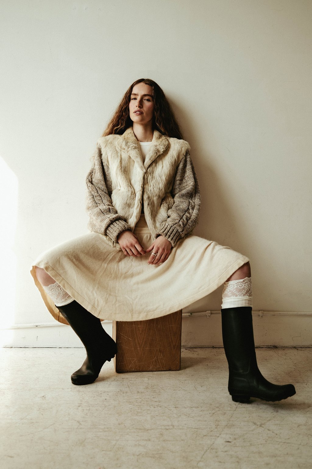 Woman leaning against a wall in a fur-collared knit jacket, cream midi skirt, and black knee-high boots