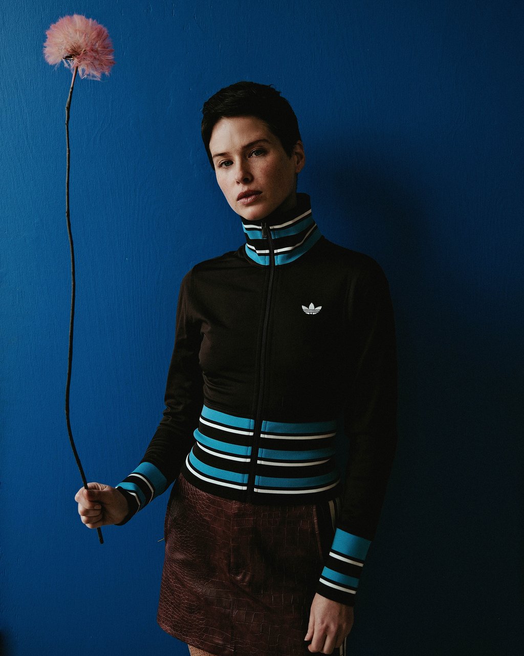 Short-haired person in a black Adidas zip jacket holding a pink fluffy flower against a deep blue wall