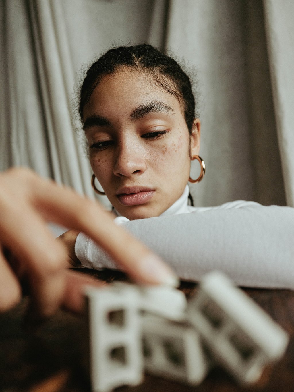 Close-up portrait of a young person with braided hair, freckles, gold hoop earrings, and a white turtleneck looking downward