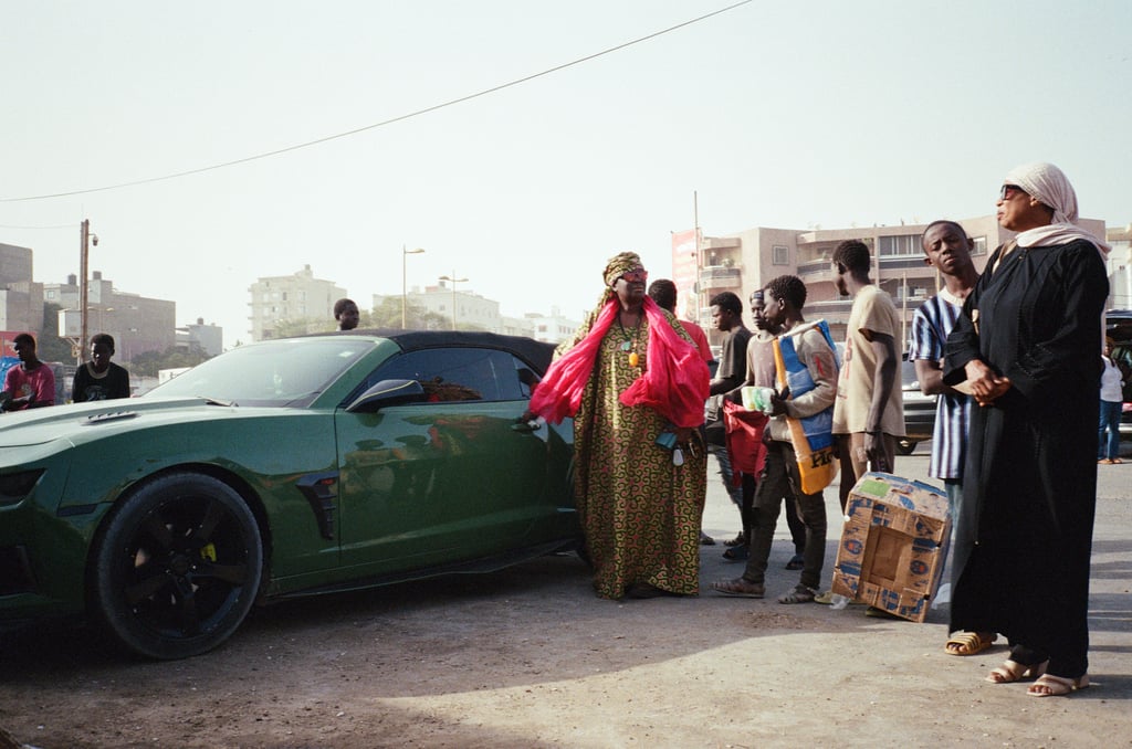 Street scene with a green convertible and a group of people gathered beside it, including a person in a patterned robe with a pink shawl