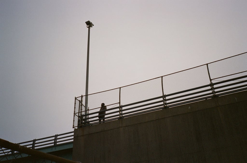 Silhouetted person standing on a raised platform behind a chain-link fence beneath a tall floodlight pole