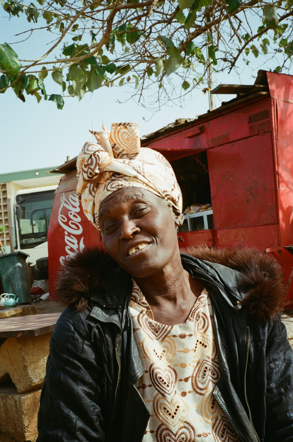 Smiling woman in a patterned headscarf and heart-motif dress with a fur-trimmed jacket near a red Coca-Cola kiosk