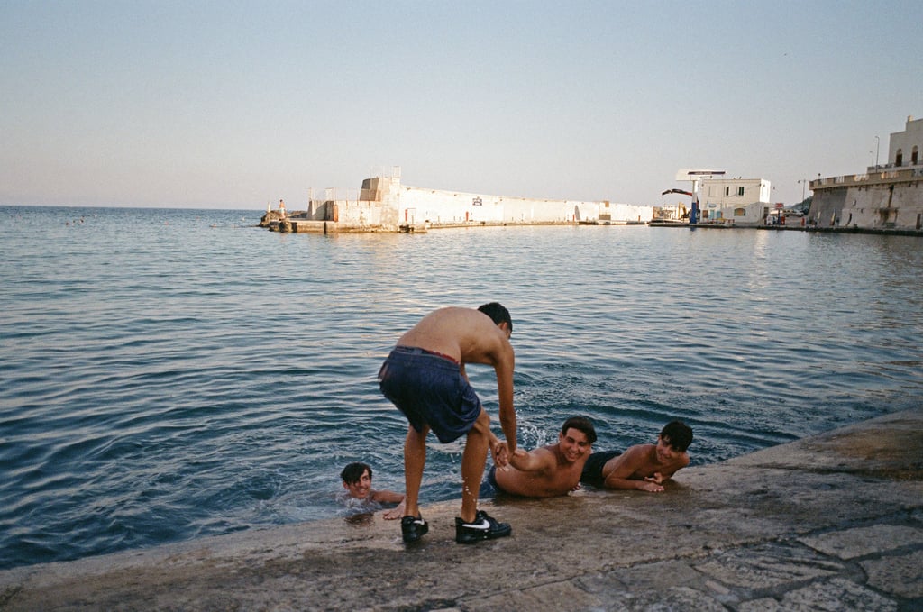 Four boys at a rocky waterfront, three in the water and one helping another climb onto the dock