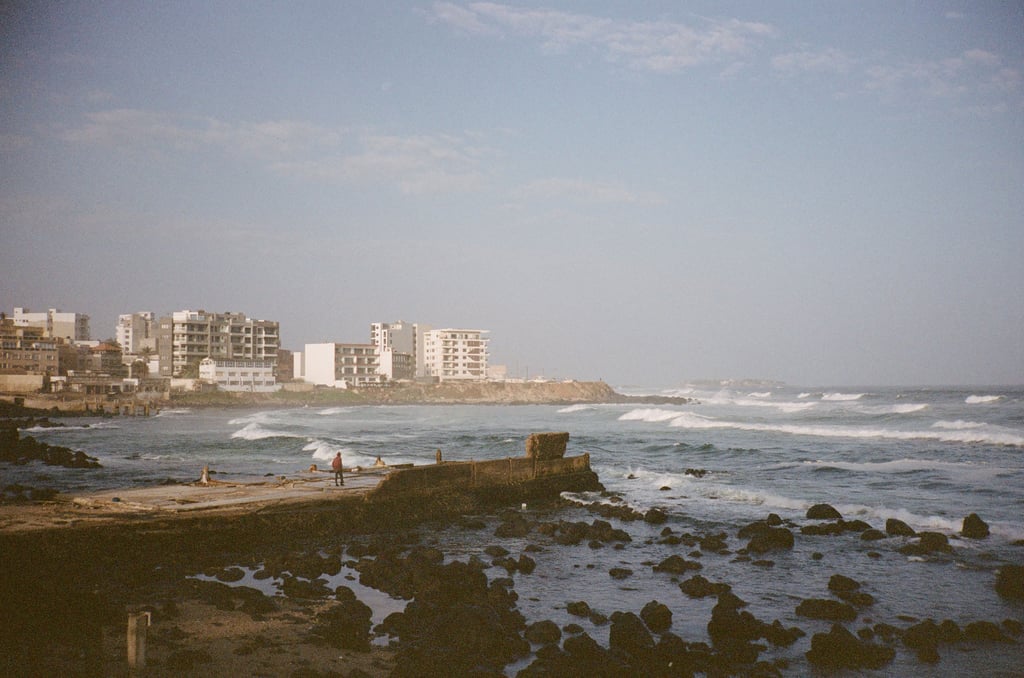 Seaside scene with low-rise apartments on a rocky headland, choppy waves, and a person standing on a concrete pier