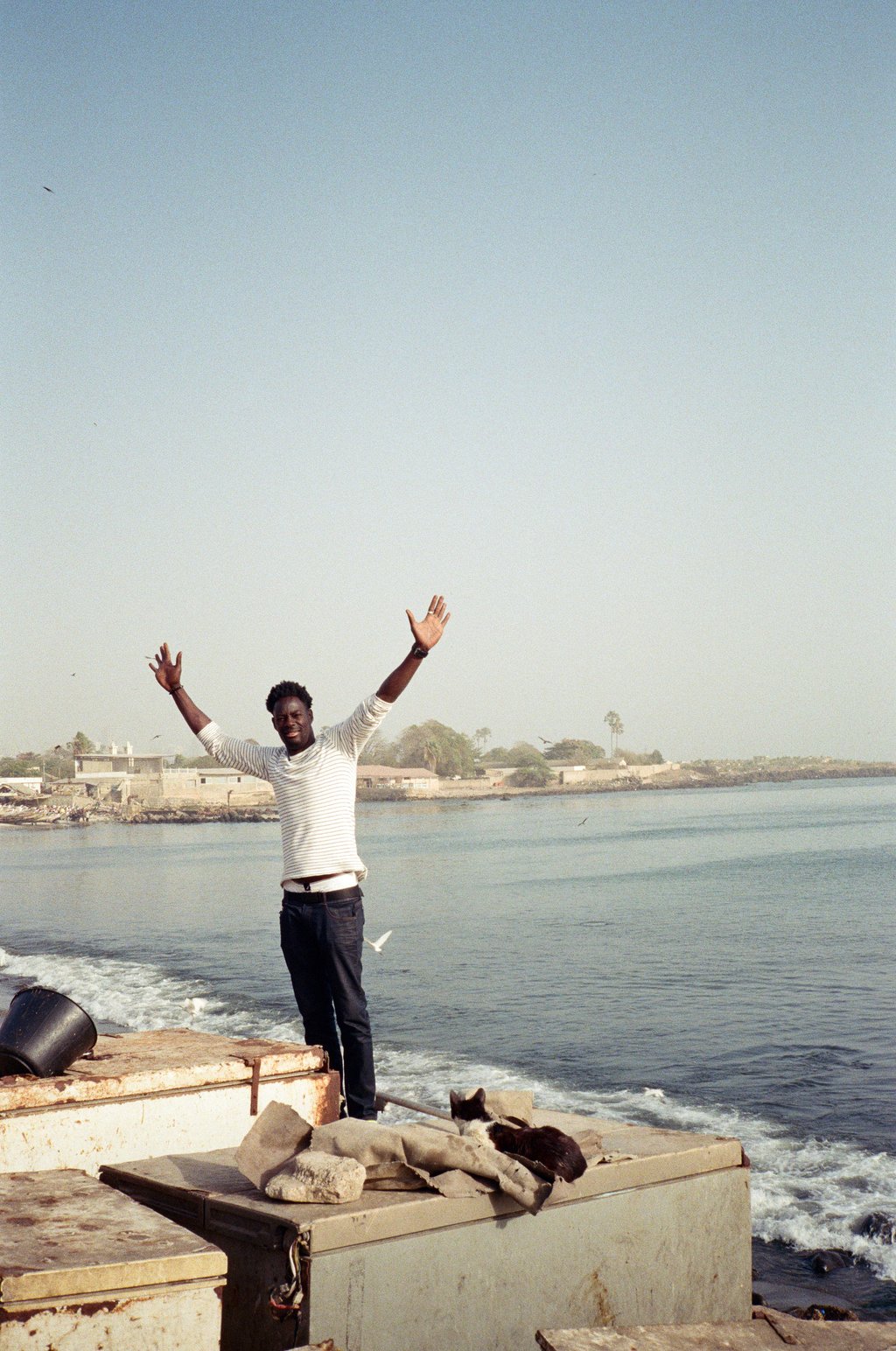 Man standing at the edge of a waterfront pier with both arms raised, a cat curled on a crate in the foreground