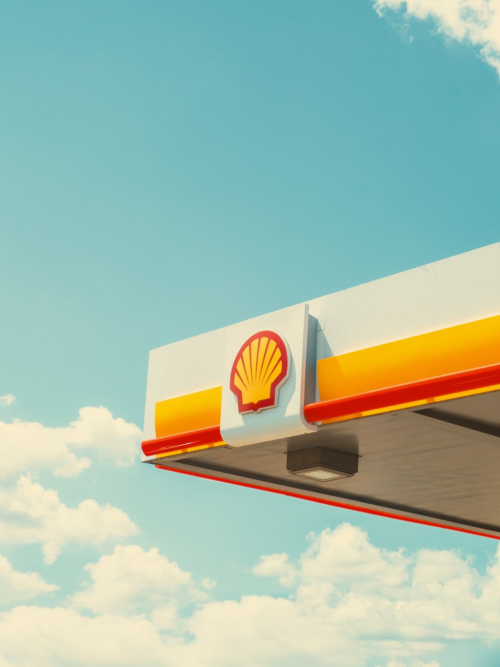 Shell gas station canopy showing the red-and-yellow Shell logo against a clear blue sky with scattered clouds