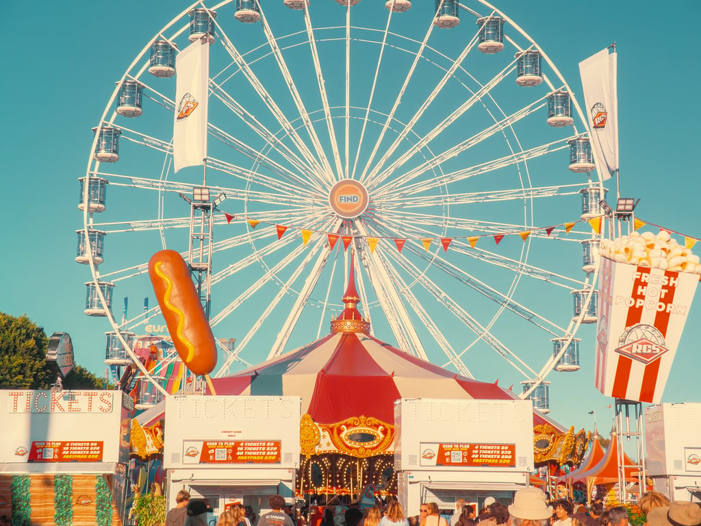 Daytime carnival scene with a large Ferris wheel, red-and-white striped tent, ticket booth, and a Fresh Hot Popcorn sign
